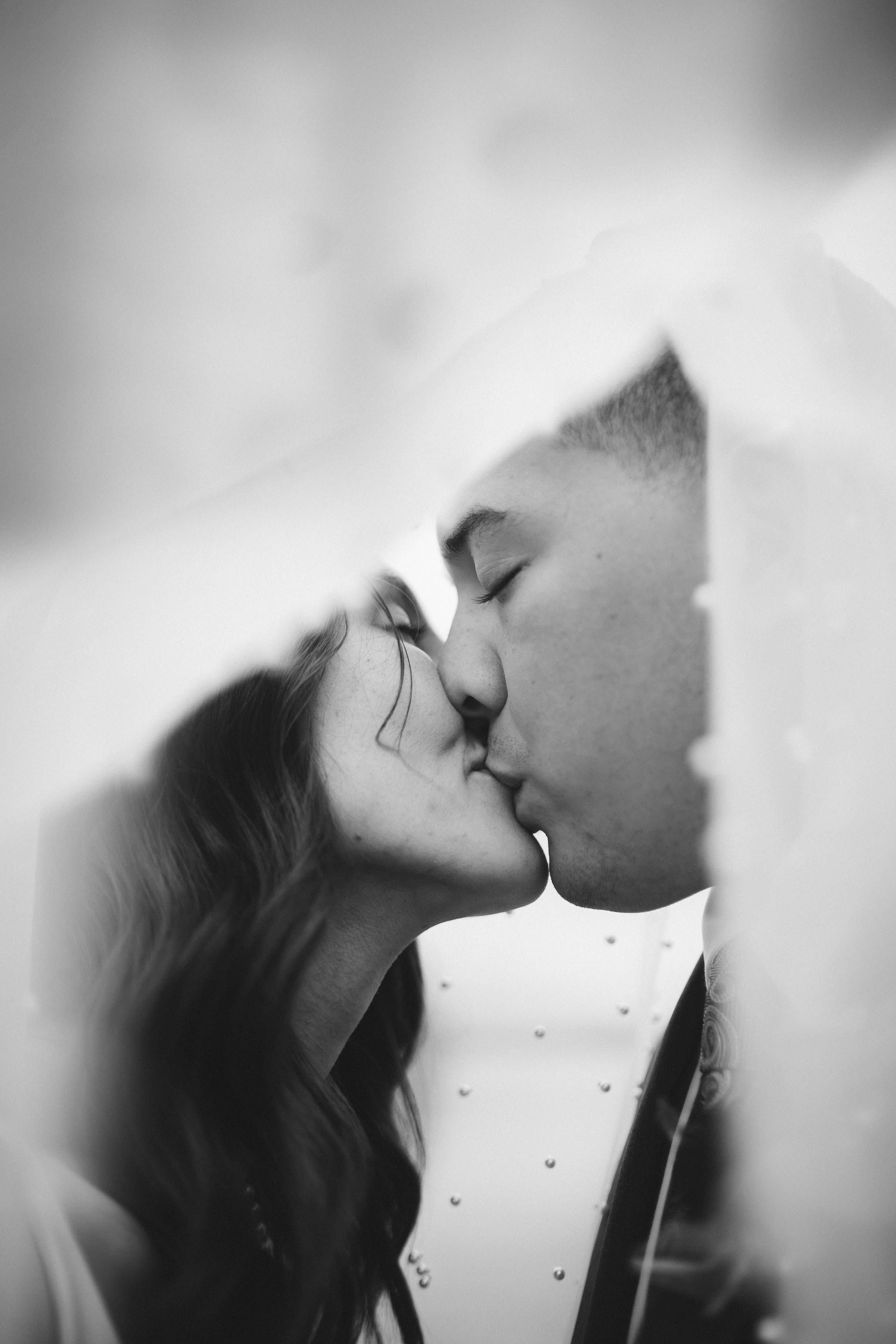 Black and white photo of a bride and groom sharing a romantic kiss beneath a flowing wedding veil.
