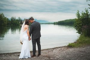 Bride and groom standing by a serene lake shore, sharing an intimate moment with veil flowing.