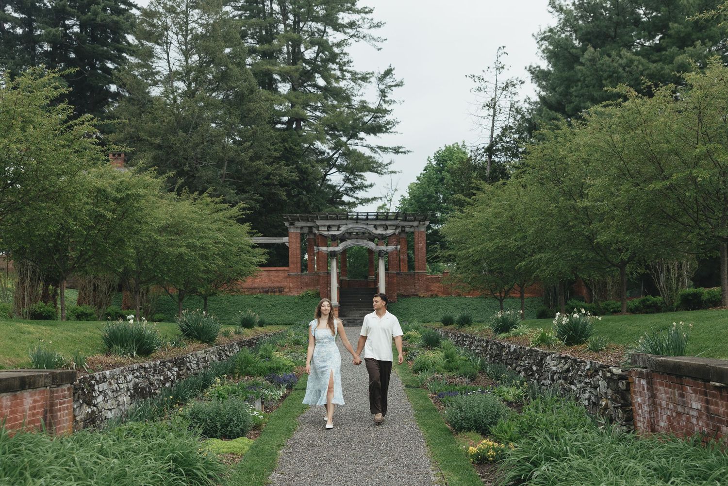 Couple walking hand-in-hand through a lush garden pathway with brick walls and tall trees.