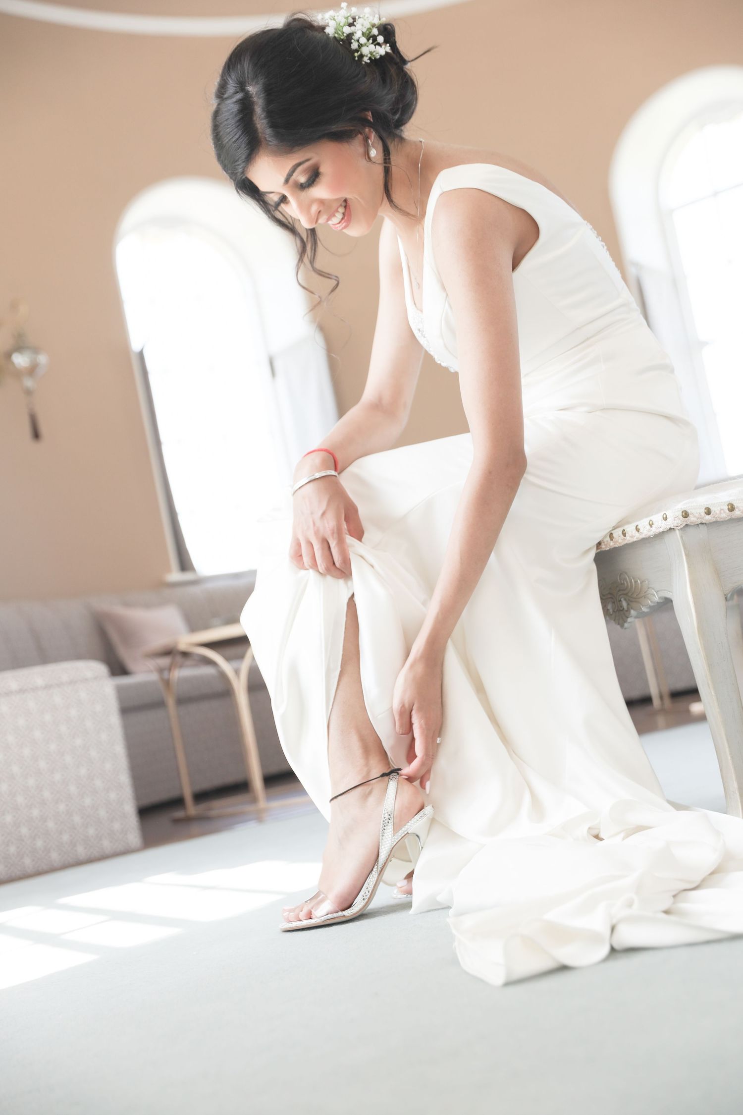 Bride in white gown fastening strappy heels while seated, with floral hair accessory, in elegant sunlit room.