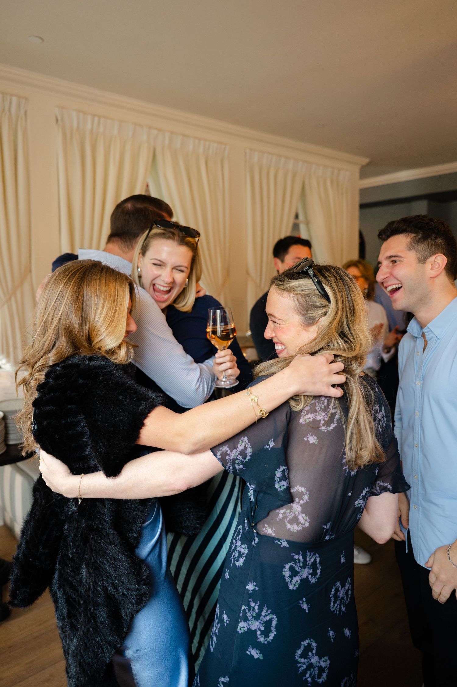 Group of friends laughing and hugging at a celebration party, woman holding champagne glass indoors.