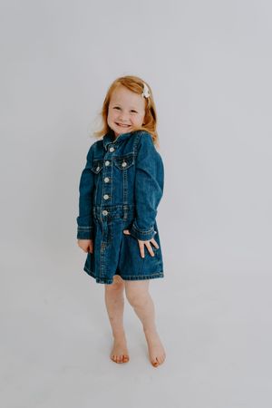 Smiling toddler girl in a denim dress with long sleeves, barefoot, posing against a light gray background.
