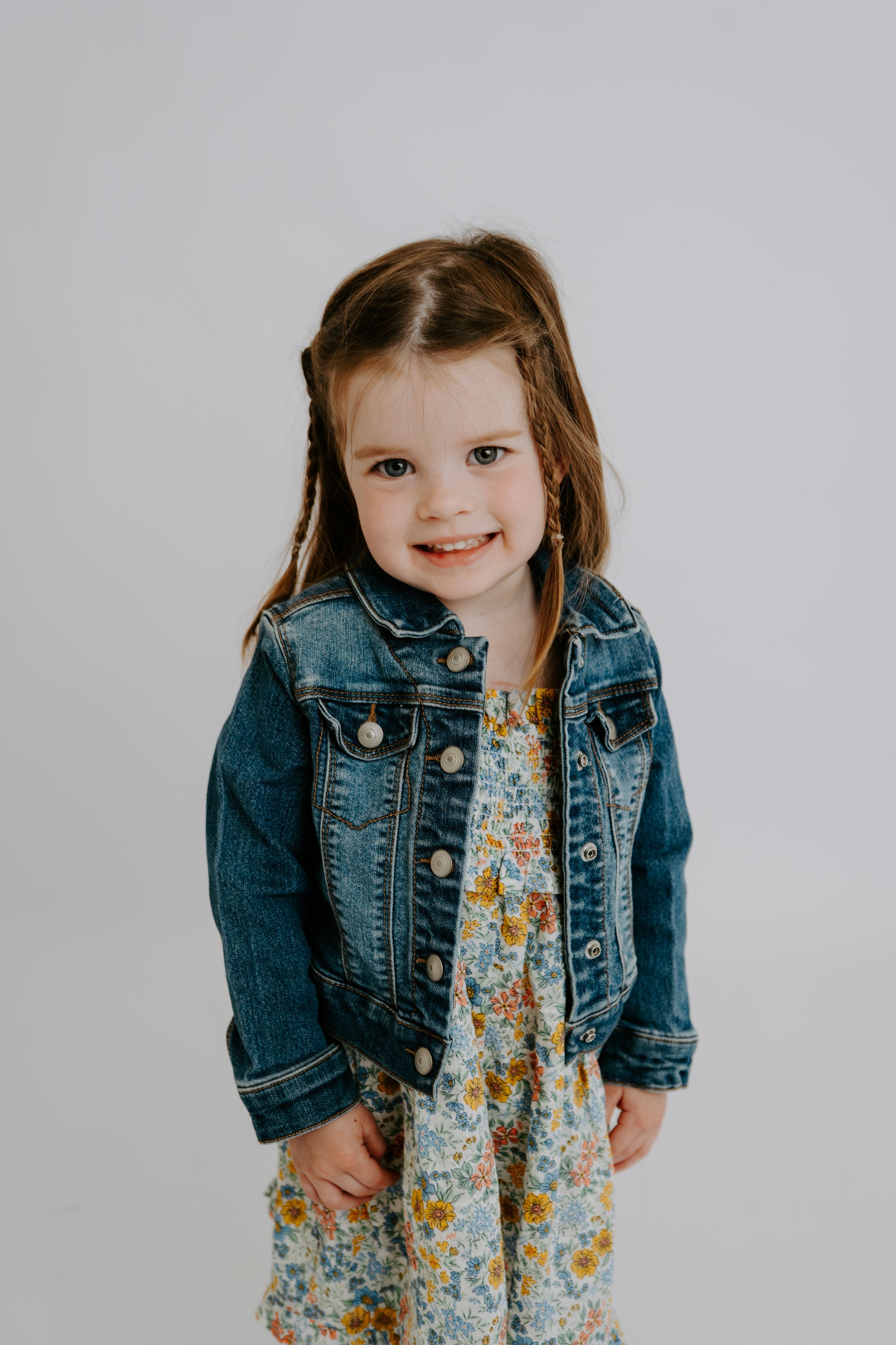A cute toddler girl smiles wearing a floral dress and denim jacket in a white studio portrait.