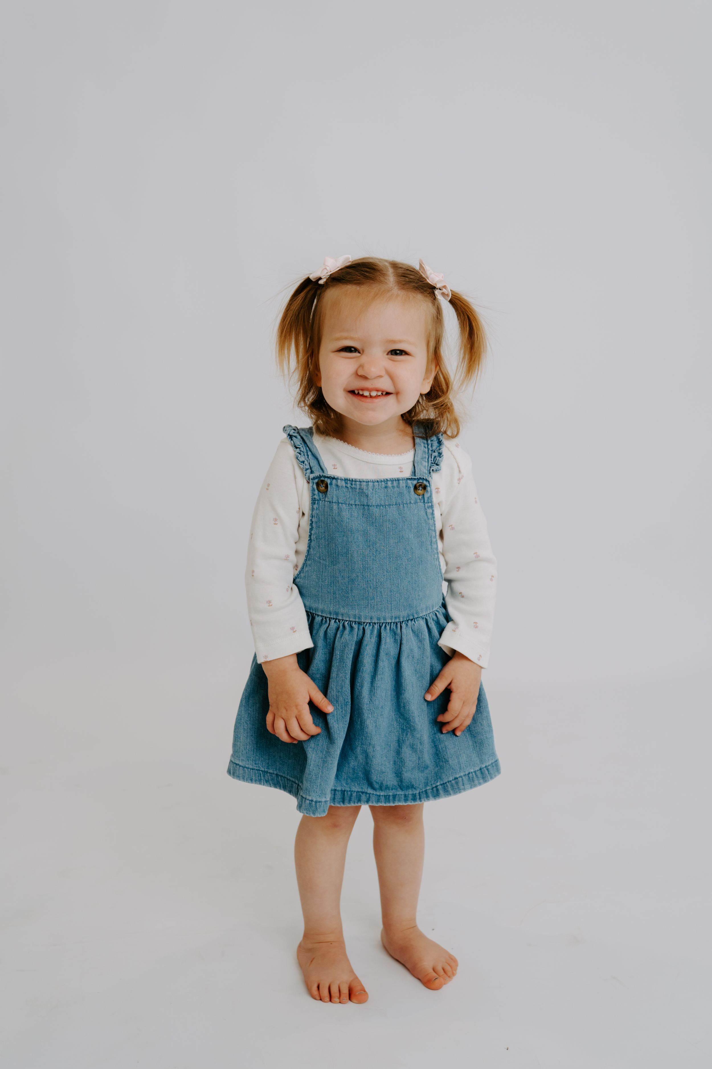 Smiling toddler girl in denim pinafore dress and white long-sleeve shirt with pigtails, standing barefoot on white background.
