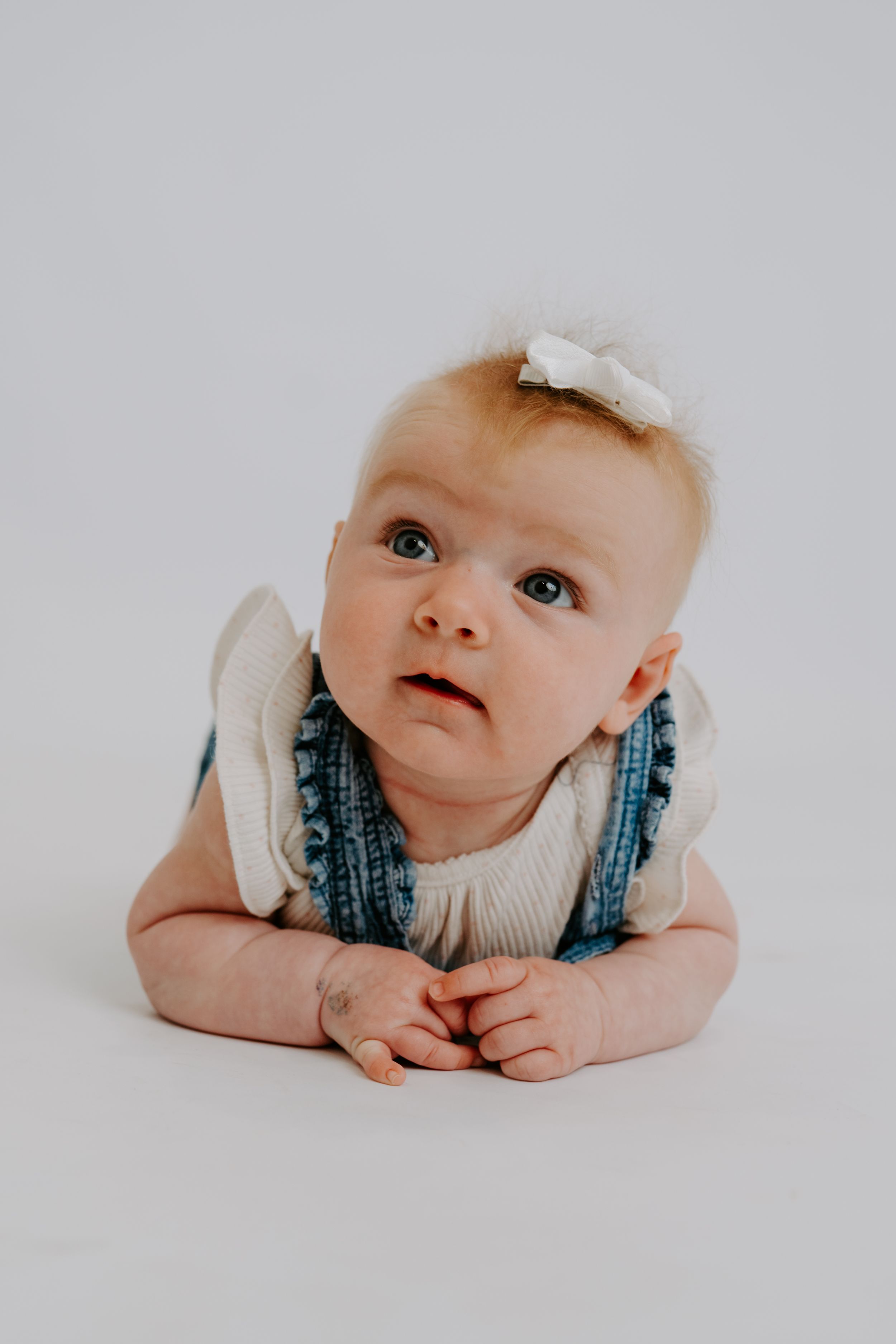 Adorable baby girl in denim overalls and white bow headband lying on tummy, looking up curiously against white background.