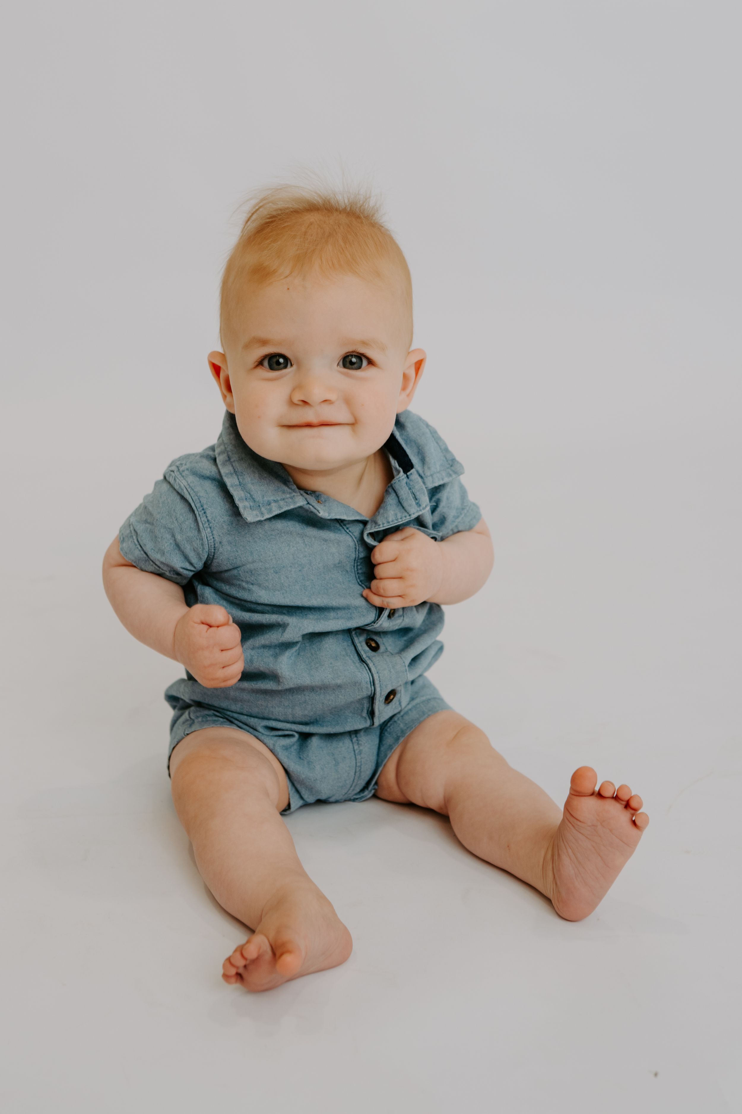 Adorable baby boy in a blue denim romper sitting barefoot on a white background, smiling at the camera.