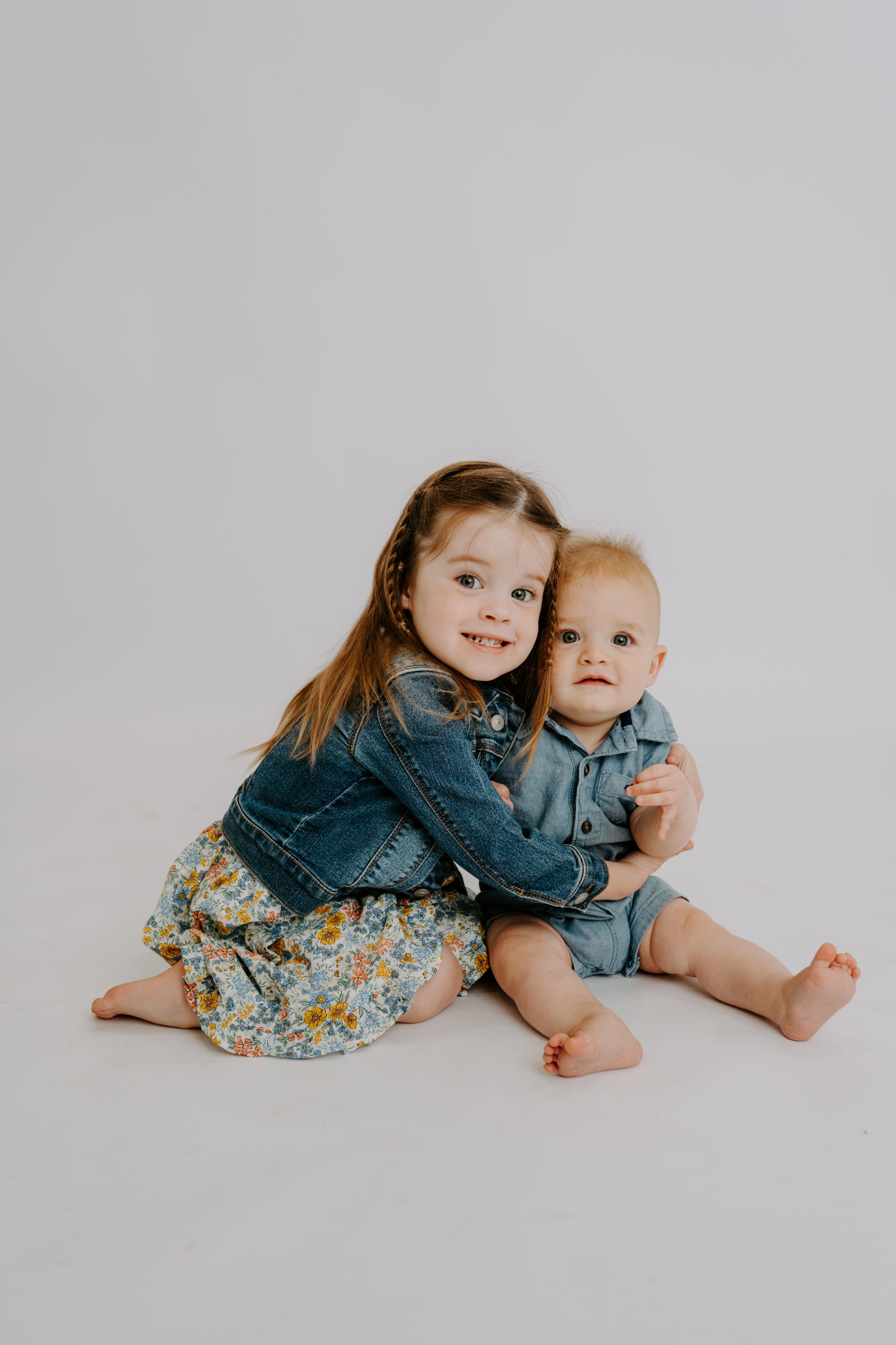 A smiling toddler girl in a denim jacket hugs a baby boy in a denim romper on a white studio background.