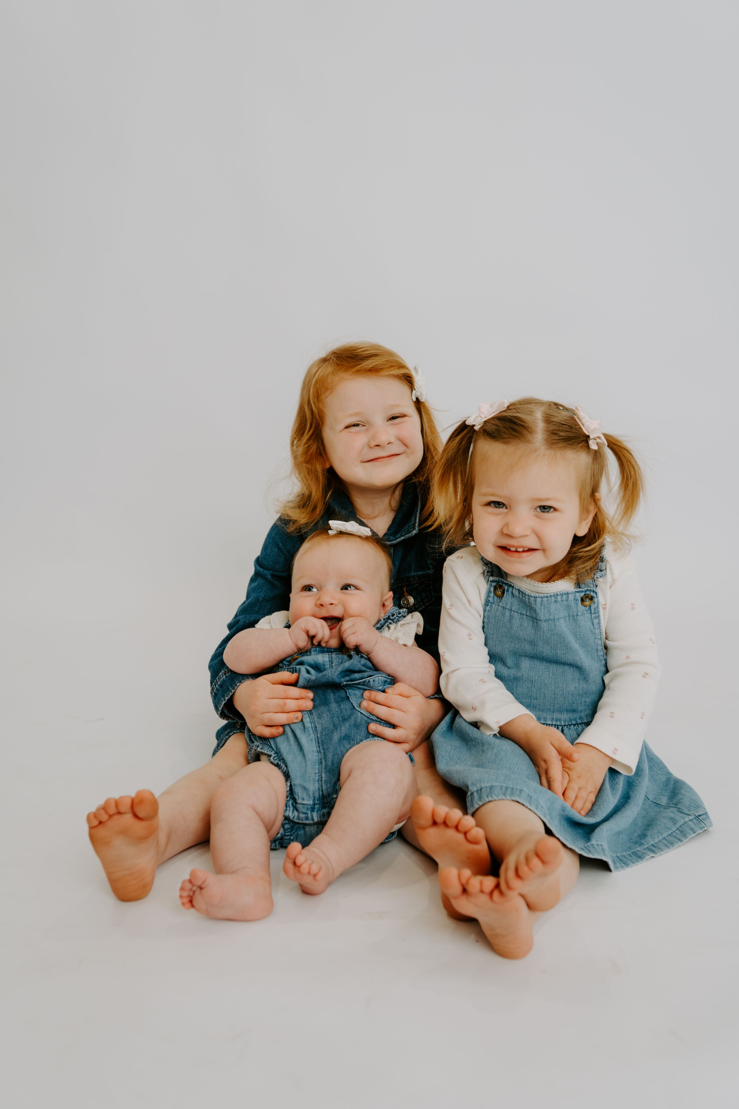 Three young siblings in denim outfits sitting together barefoot on a white background, smiling warmly.