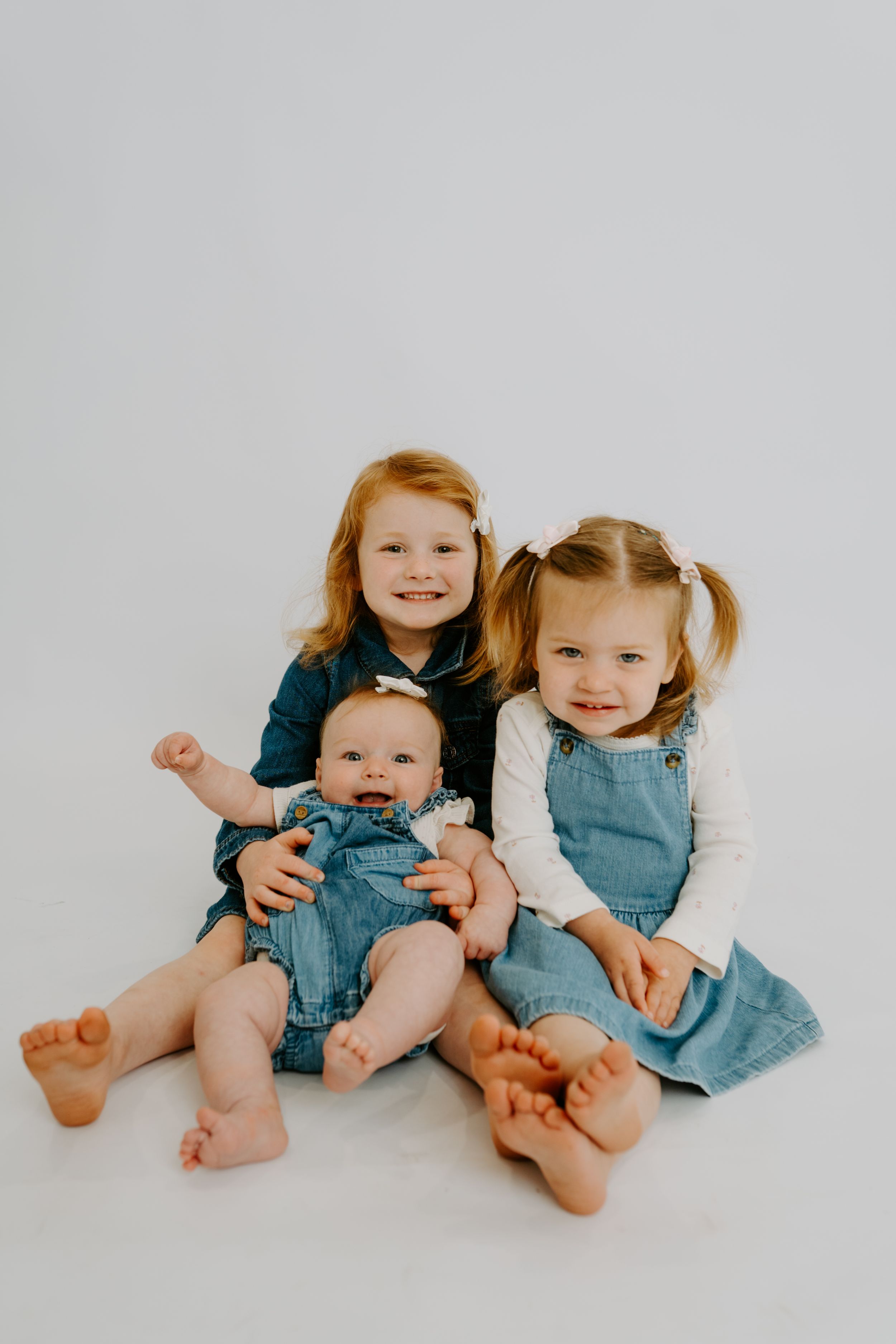 Three young siblings in denim outfits pose together on white background, two toddler girls holding a smiling baby.