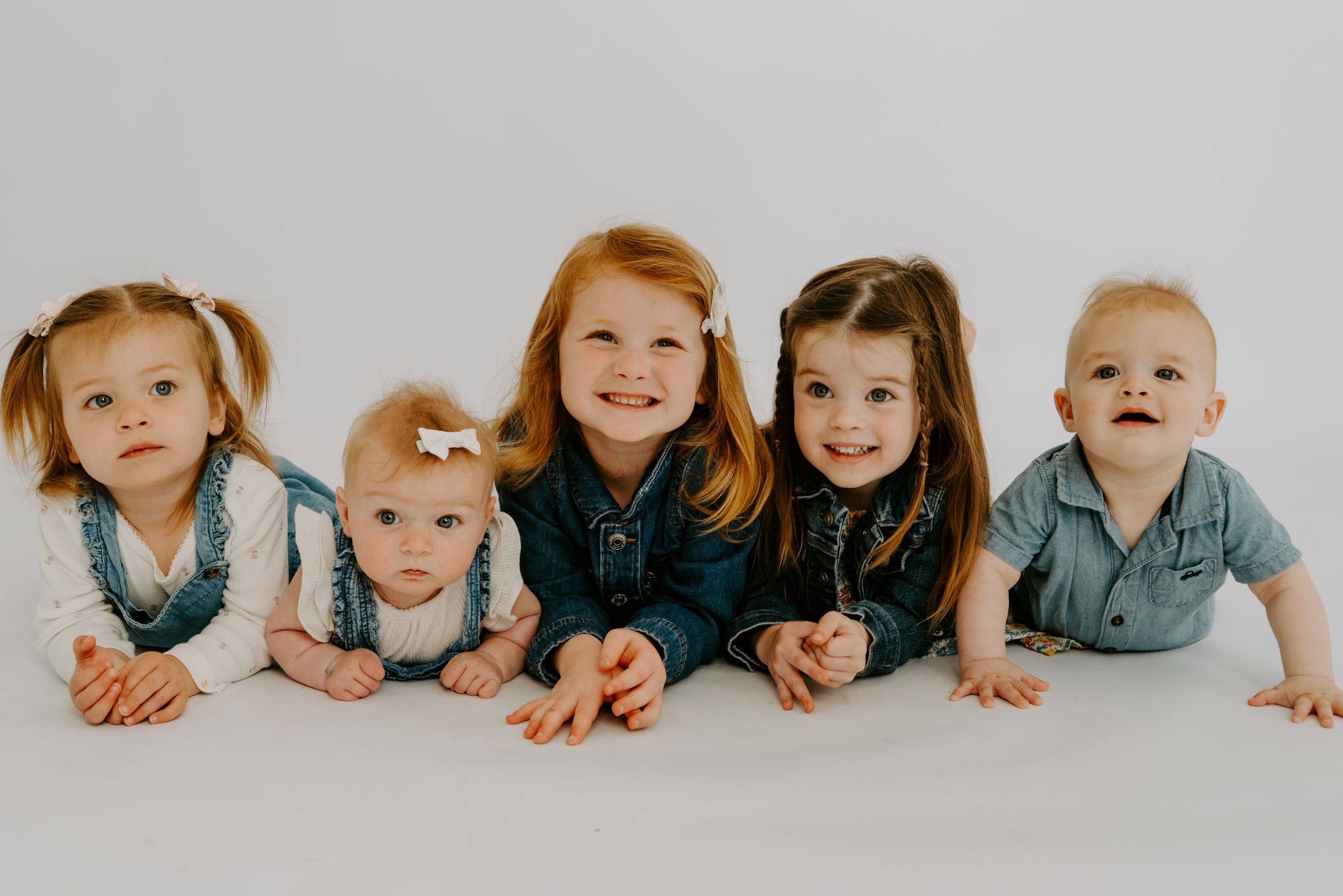 Five young children pose lying down in a row, smiling at the camera against a white studio background.