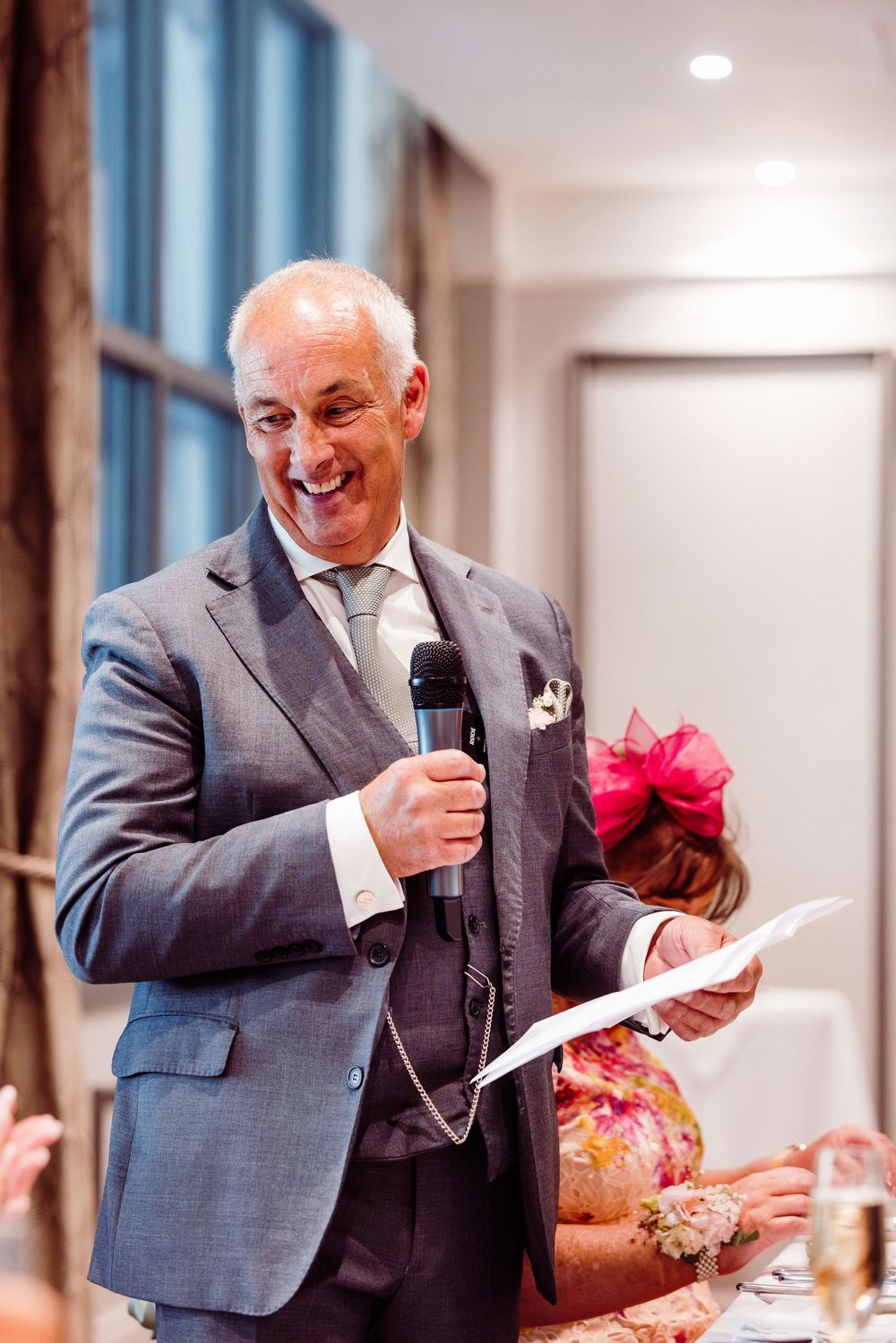 Smiling older man in gray suit giving a speech at a wedding reception while holding a microphone and papers.