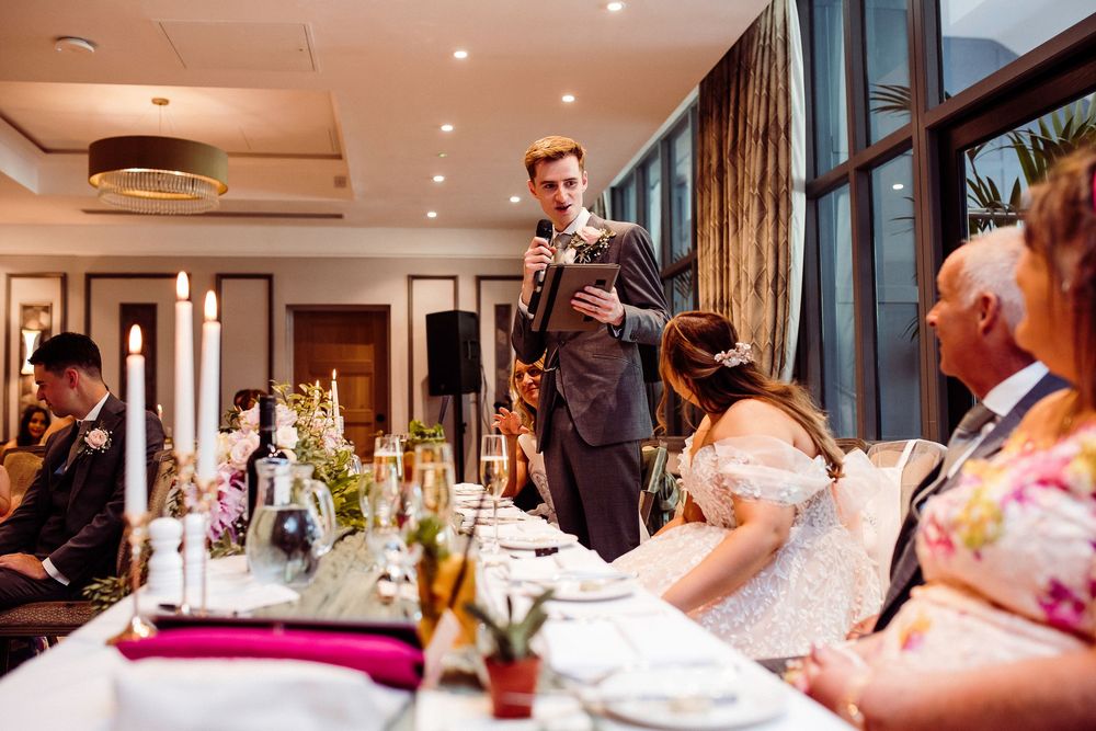 Groom giving a wedding speech at an elegant reception dinner table with candles and floral centerpieces.