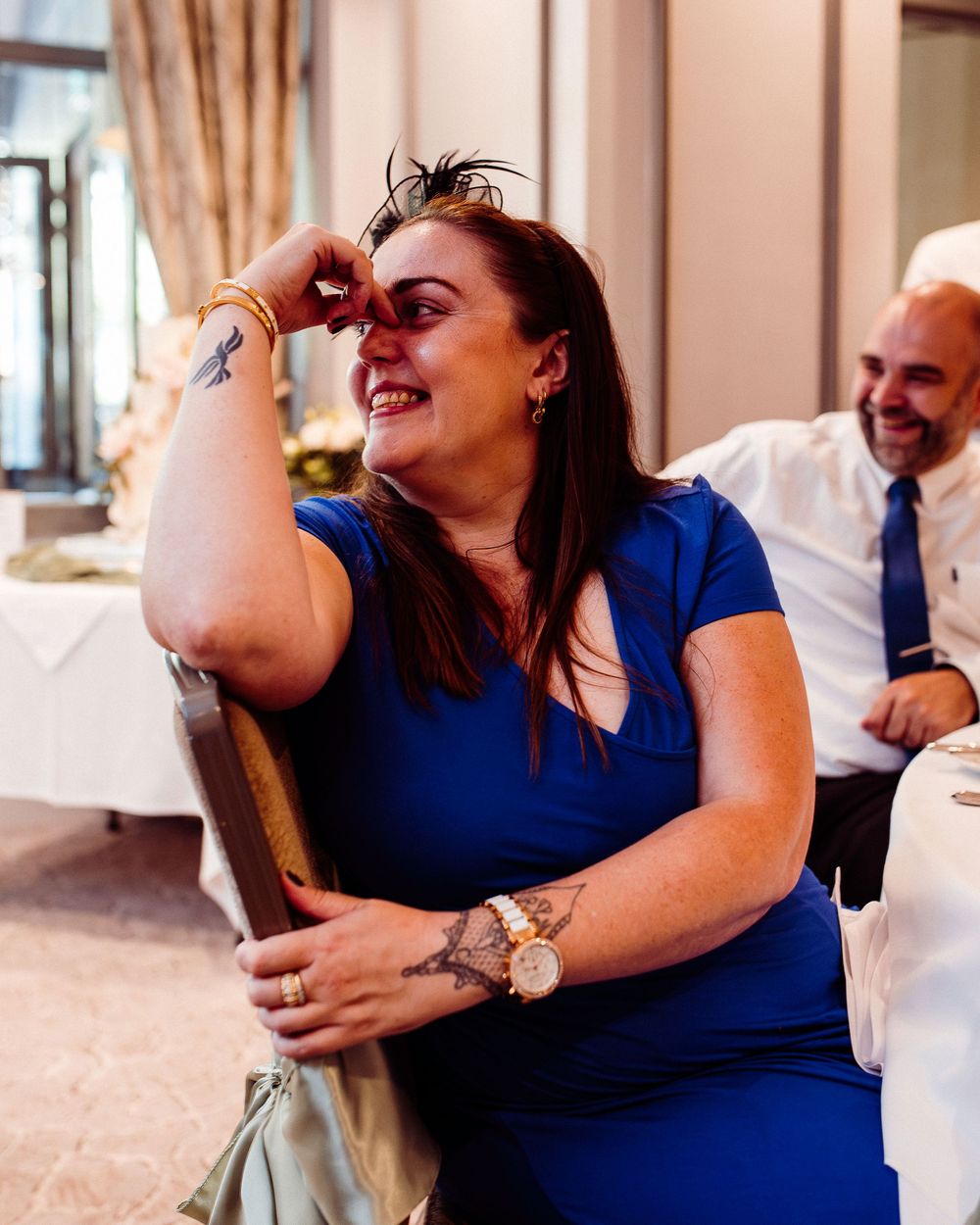 A laughing woman in a blue dress with tattoos sits at a wedding reception, holding a chair back, with a smiling man behind her.