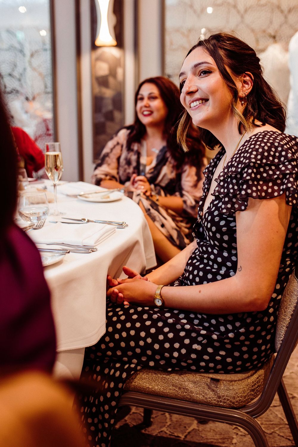 Women in polka dot dresses smiling and laughing at an elegant restaurant dinner table with champagne.