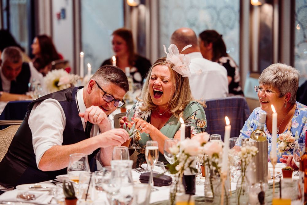 Guests laughing and enjoying a magic trick at a wedding reception dinner table with candles and flowers.