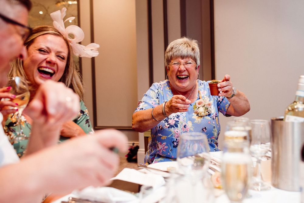 Two women laughing joyfully at a wedding reception dinner table, wearing floral outfits and hair accessories.
