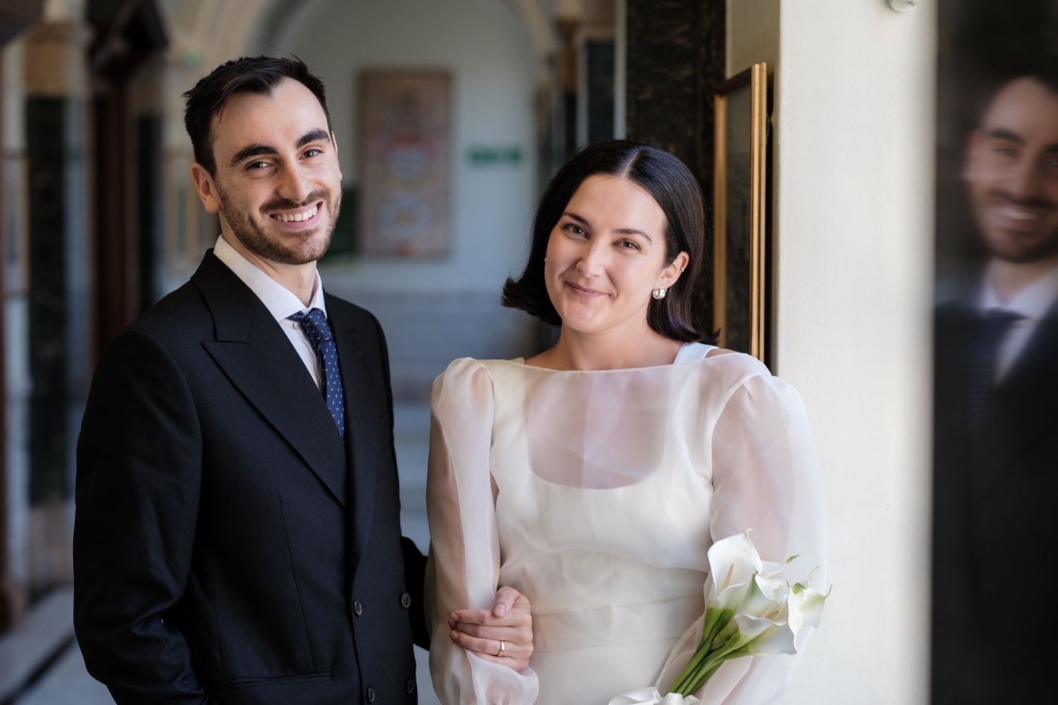 Smiling couple in wedding attire, groom in black suit, bride in white gown holding calla lilies, posing indoors.