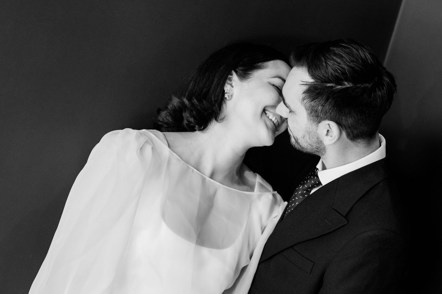 Black and white photo of a smiling couple in formal wear sharing an intimate moment, touching foreheads together.