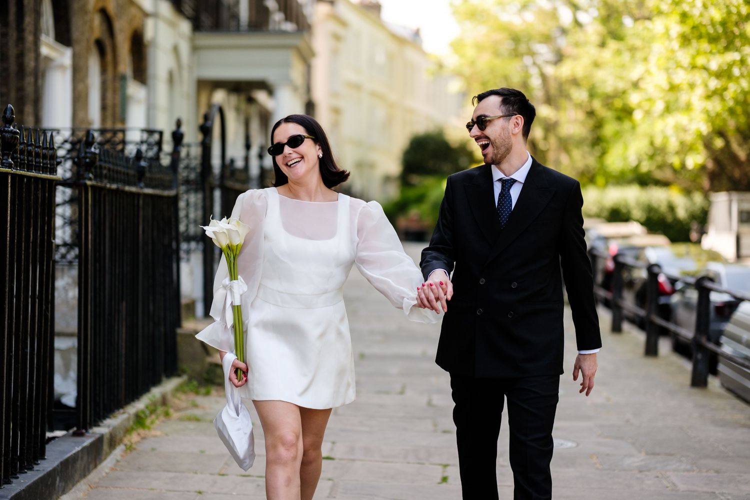 Smiling couple in sunglasses walking hand-in-hand on a sunny sidewalk, bride in white mini dress holding roses, groom in black suit.