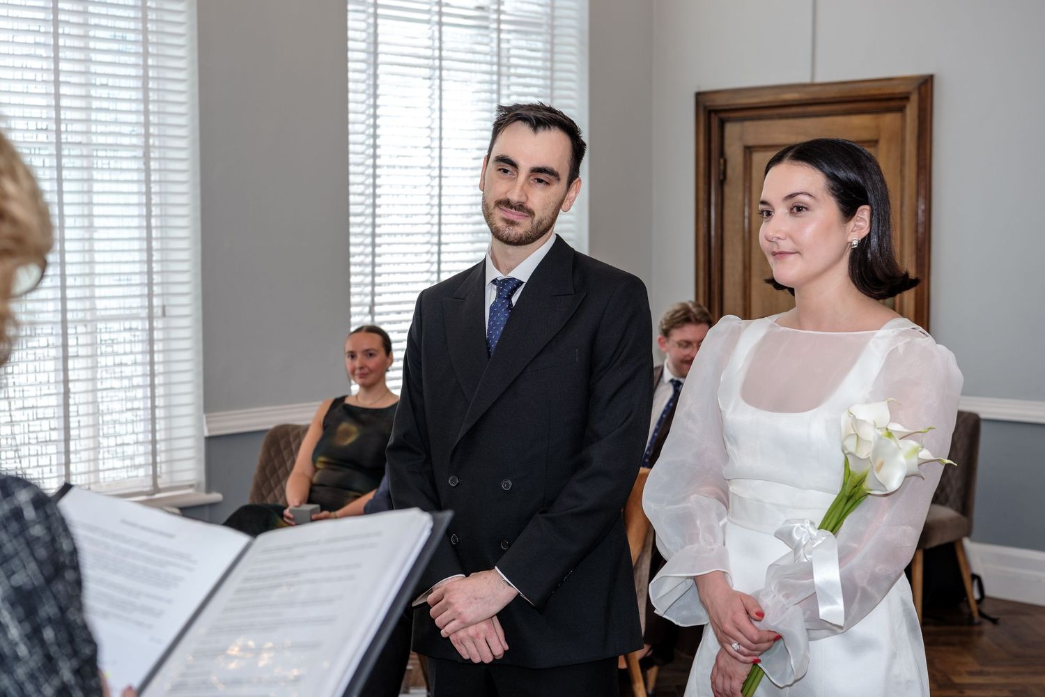 Couple exchanging wedding vows at a civil ceremony, groom in black suit, bride holding white calla lilies.