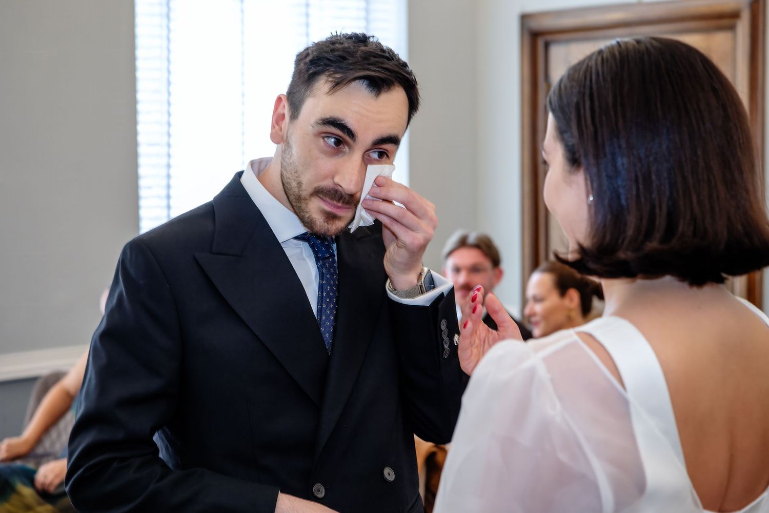Emotional groom wiping tears at wedding ceremony while bride stands before him in white dress.