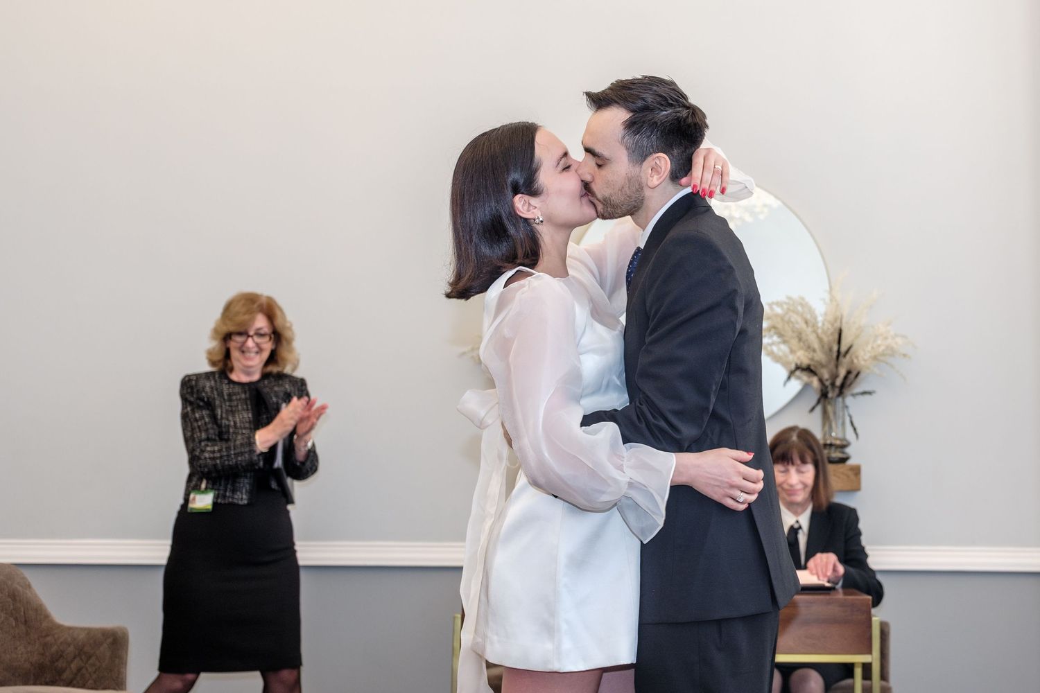 Couple shares first kiss at civil wedding ceremony while guests applaud in elegant white room.