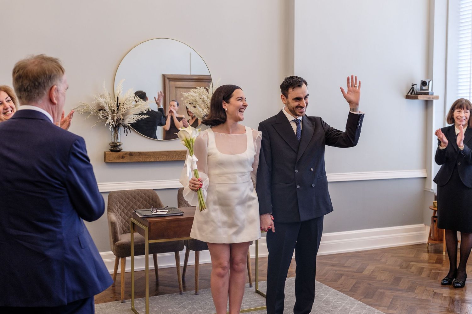 Joyful newlywed couple waving and smiling at guests during intimate wedding ceremony indoors.