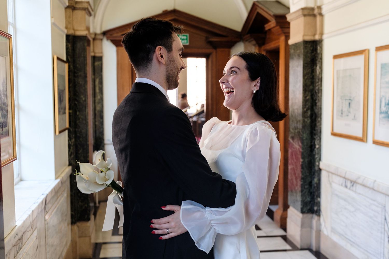 Bride and groom share a joyful embrace in an elegant hallway, bride holding white calla lilies wearing a white gown.