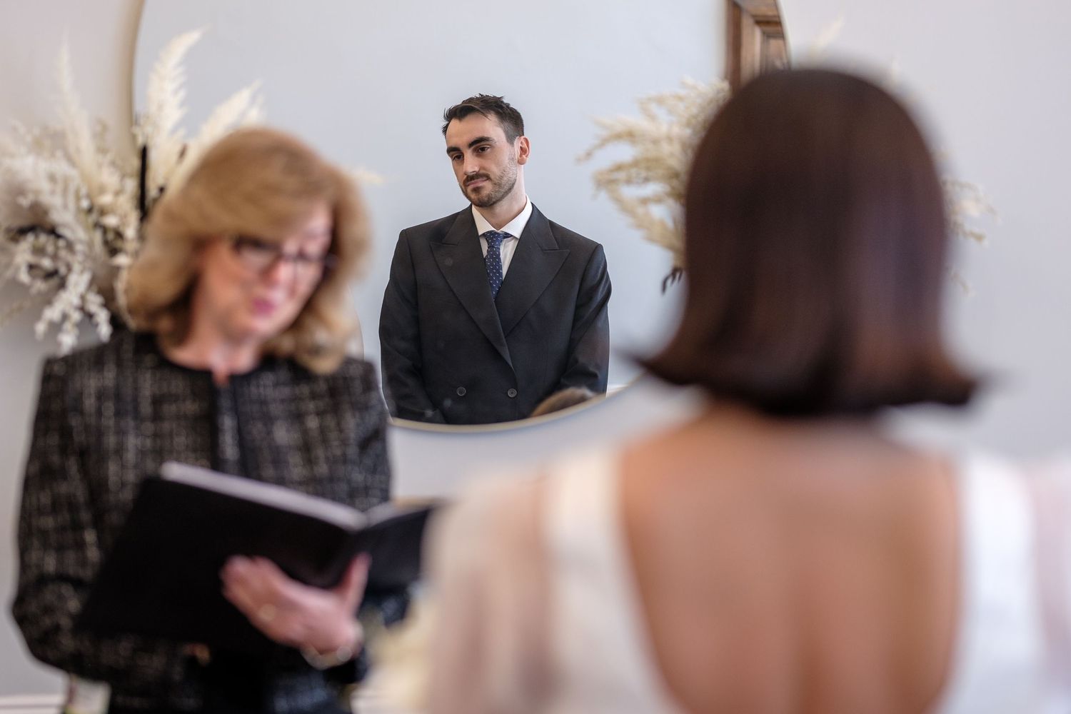 Groom in dark suit watches bride during intimate wedding ceremony with officiant reading from book.
