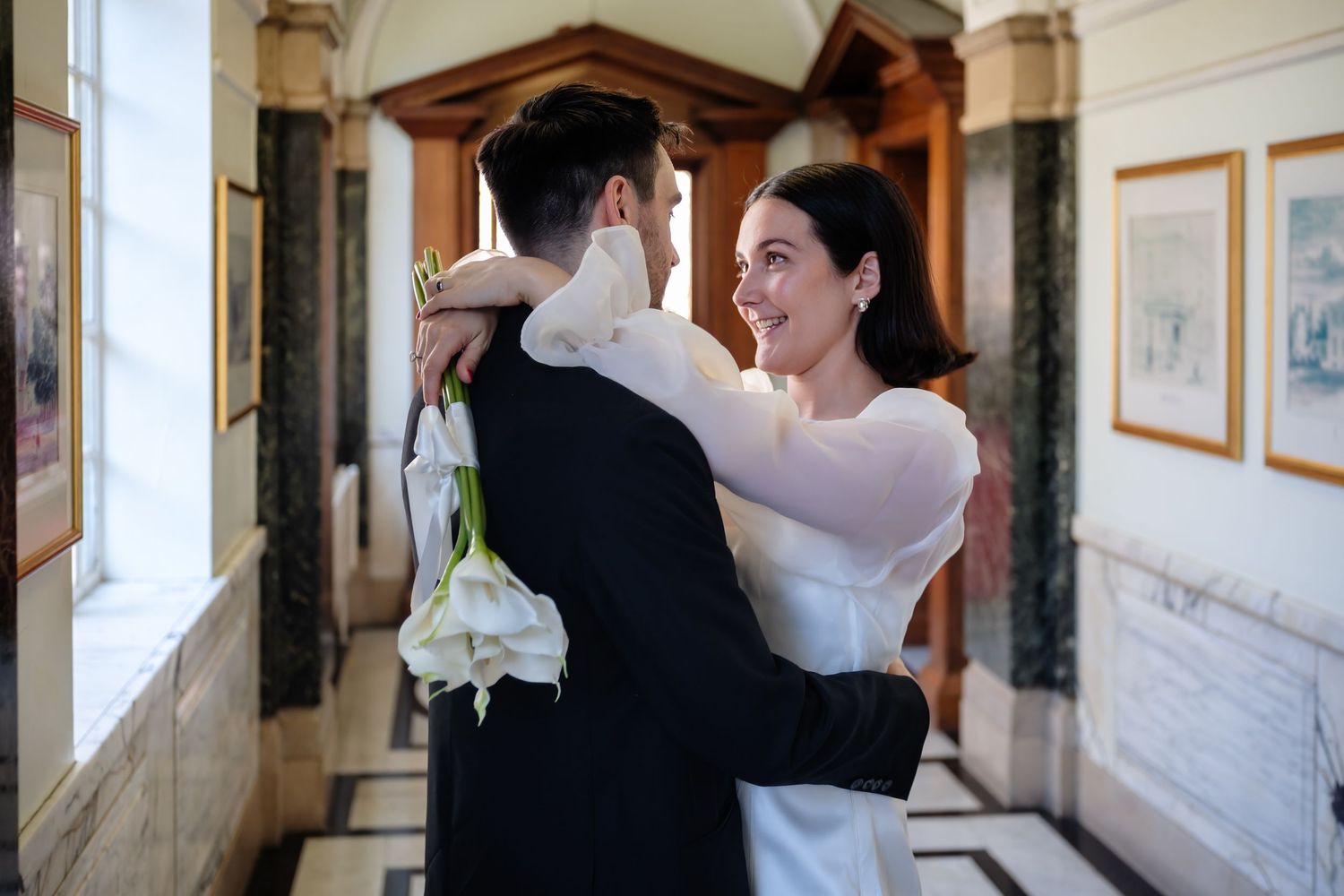 Happy bride hugs groom in a classic interior hallway, holding white calla lily bouquet in a elegant white wedding dress.