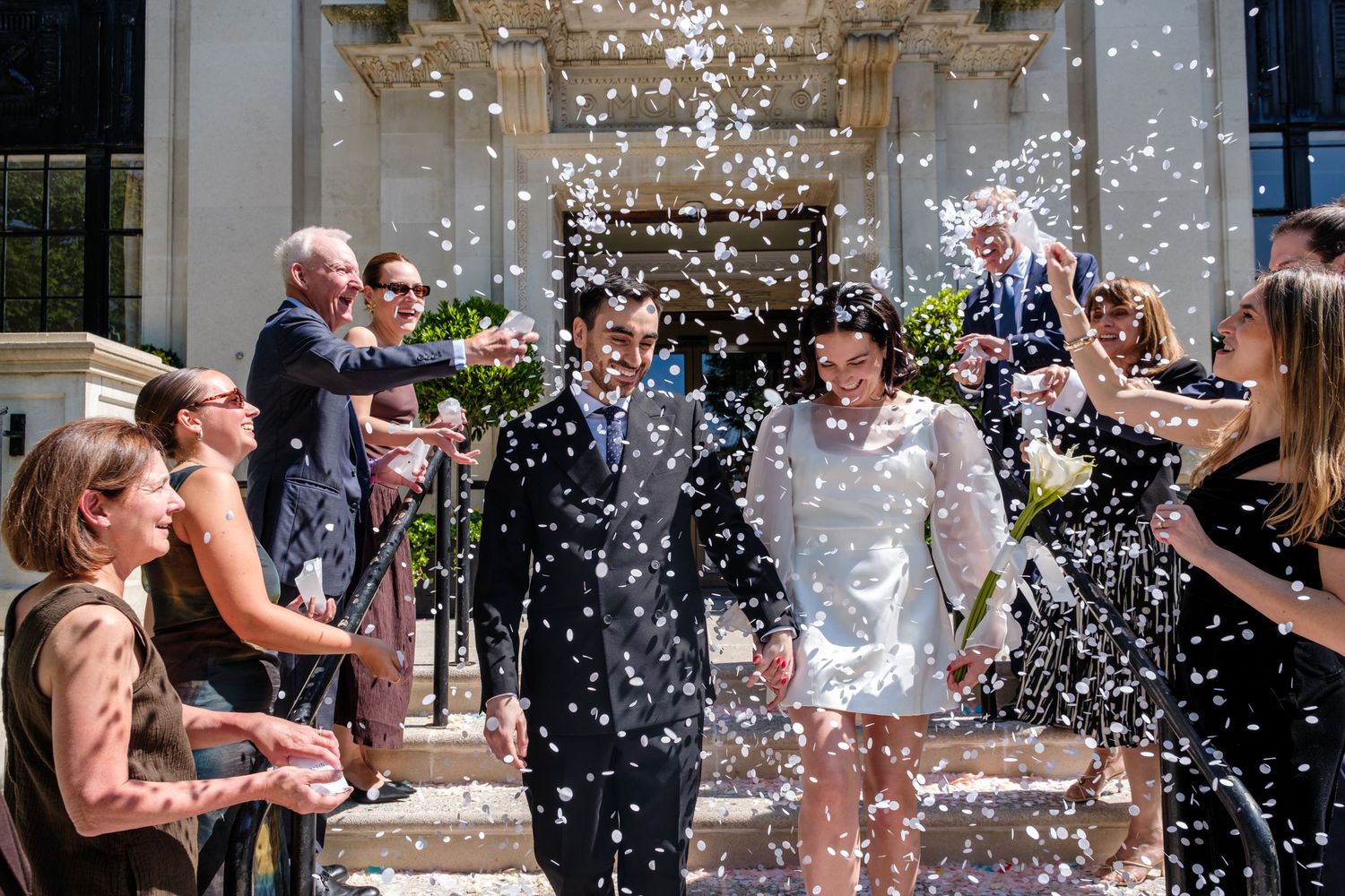 Bride and groom walk through confetti shower surrounded by cheering guests outside elegant building on sunny wedding day.