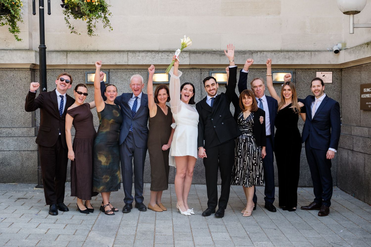 A joyful wedding group photo with eleven people raising their arms in celebration outside a stone building.