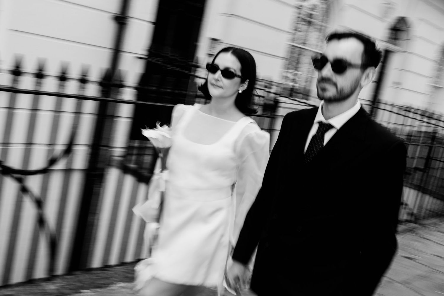 Black and white photo of a stylish couple in sunglasses walking together, woman in white dress, man in dark suit.