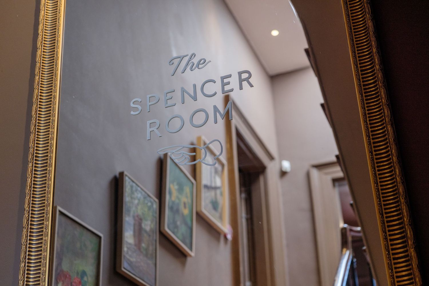Gold-framed mirror reflecting The Spencer Room sign on a gray wall with artwork in an elegant interior.