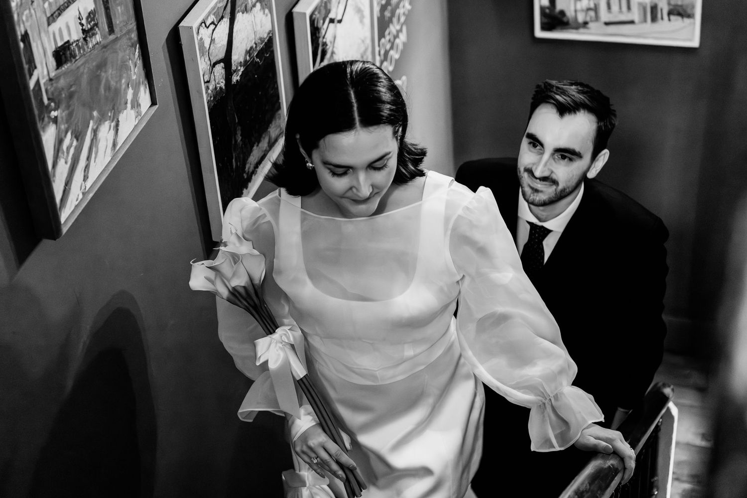 Black and white photo of a bride in a white gown holding flowers, smiling as a suited groom follows her up stairs.