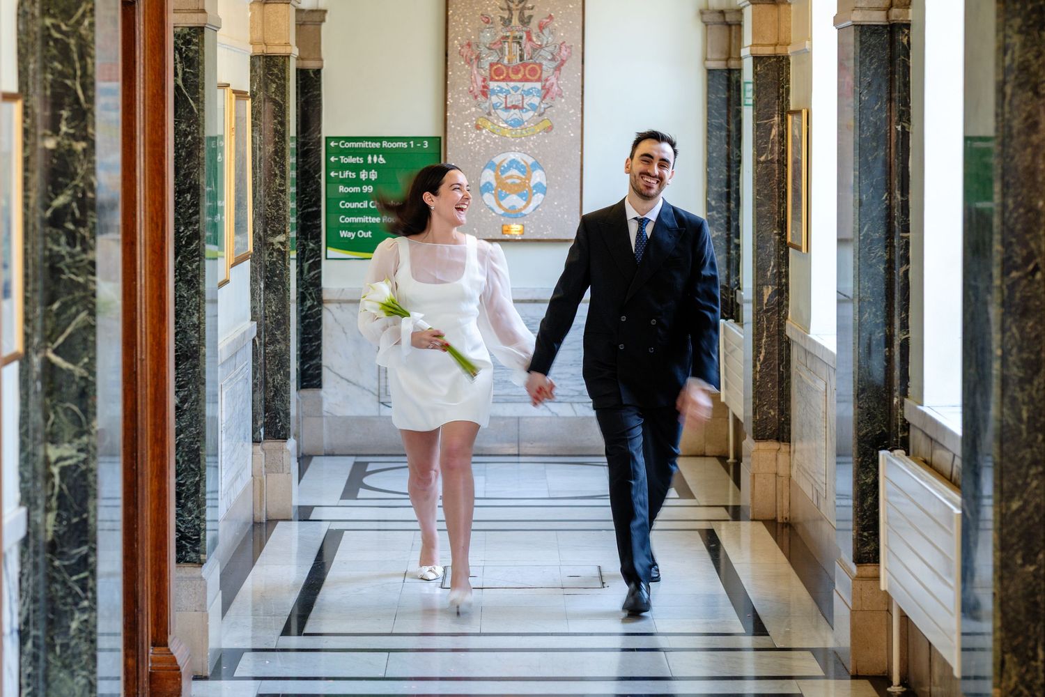 Happy newlyweds walking hand-in-hand through an elegant marble hallway, bride in white mini dress holding bouquet.