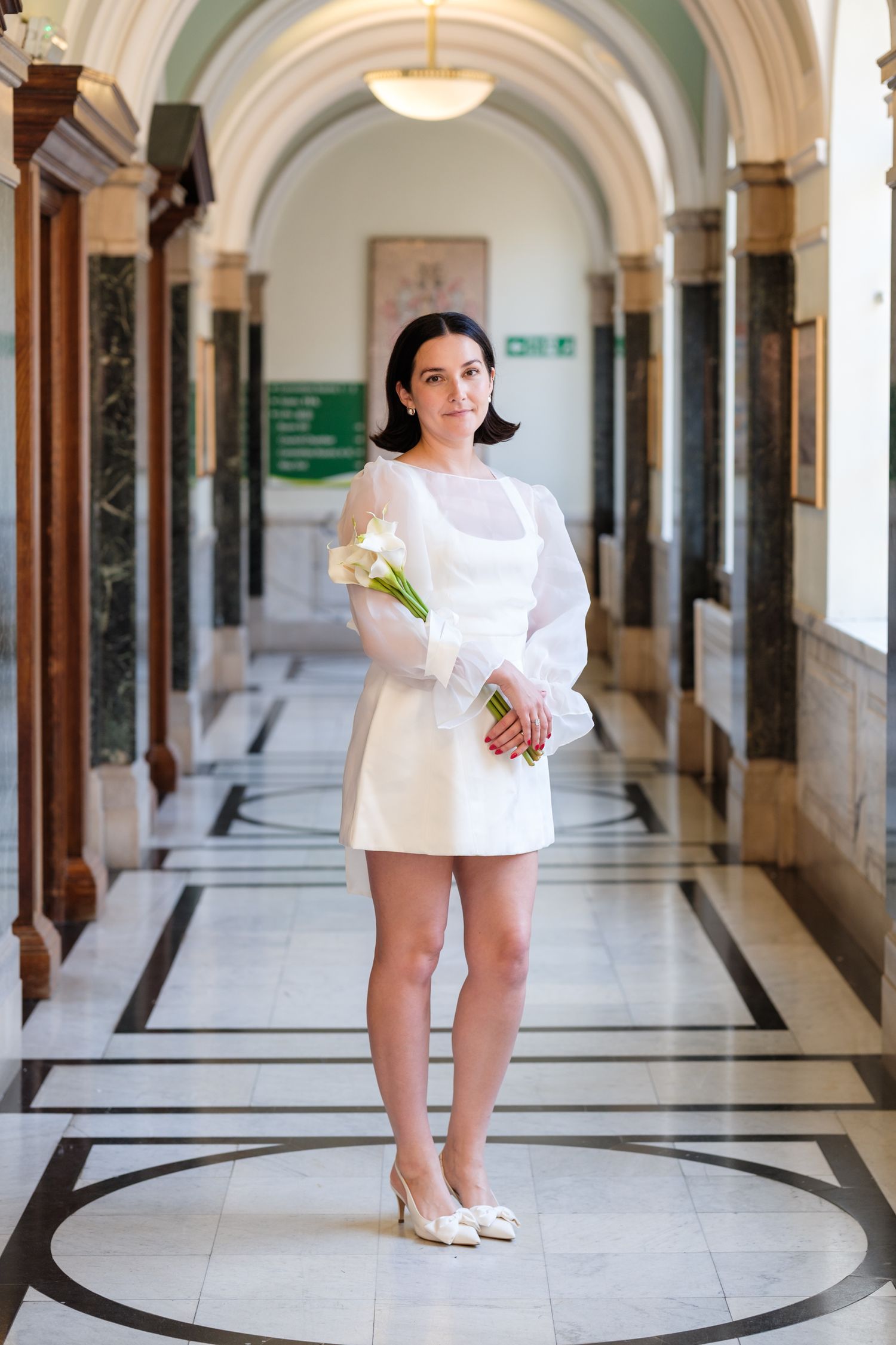 Bride in short white dress with sheer sleeves holding flowers in elegant marble hallway.