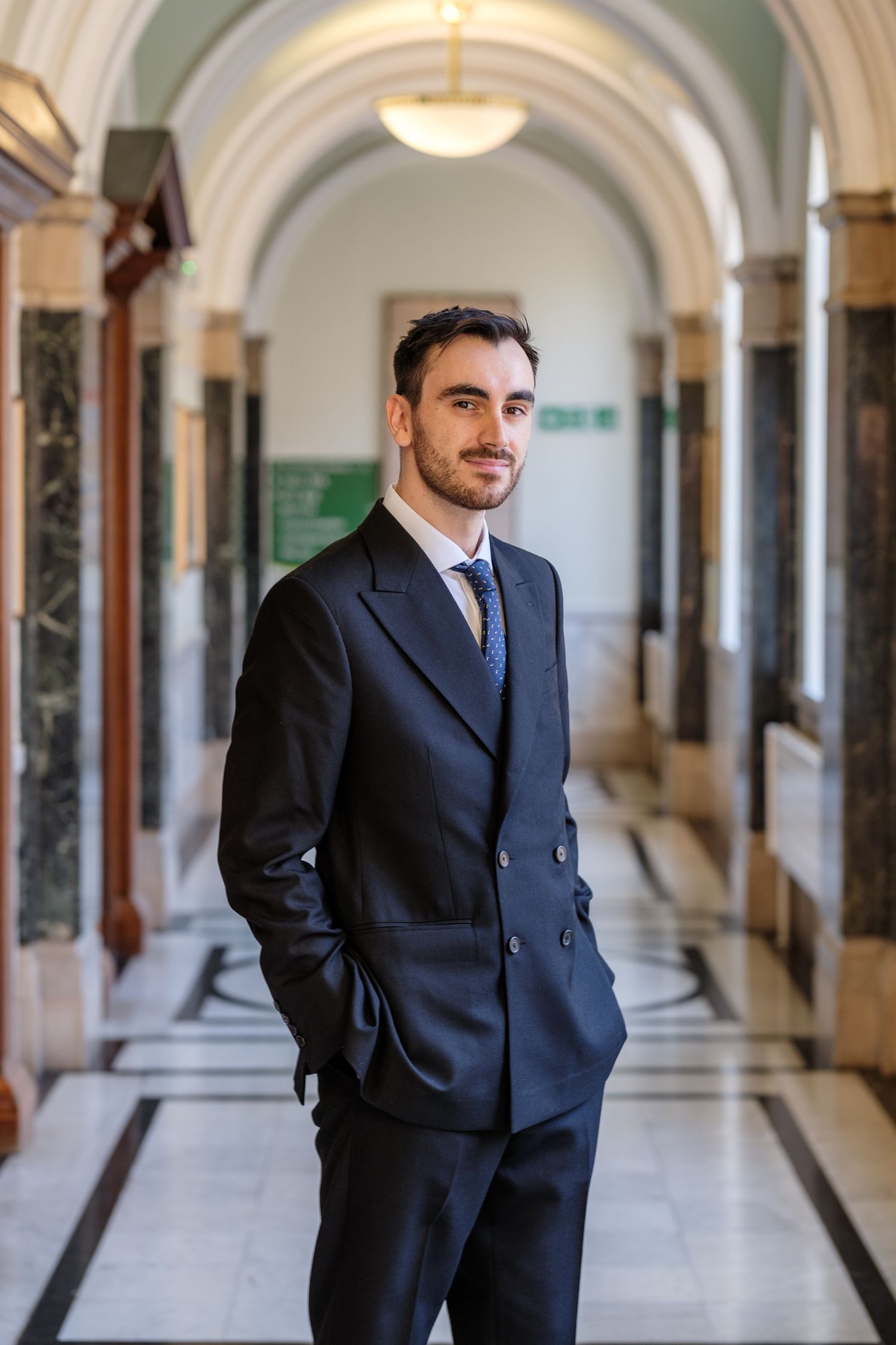Young professional man in navy suit standing confidently in elegant marble hallway with arched ceilings.