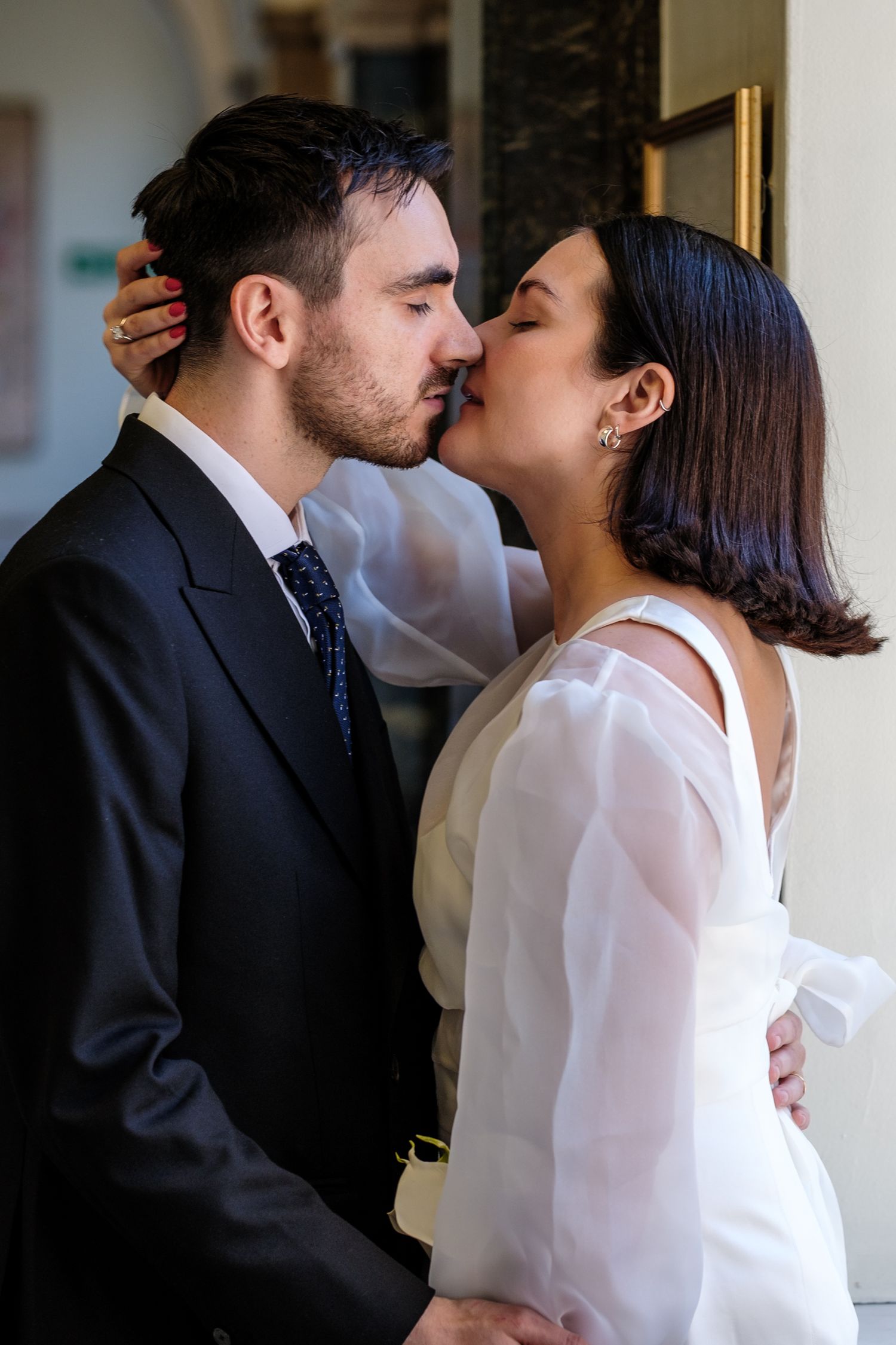 A couple shares a romantic kiss, the groom in a dark suit and the bride in a white dress with sheer sleeves.