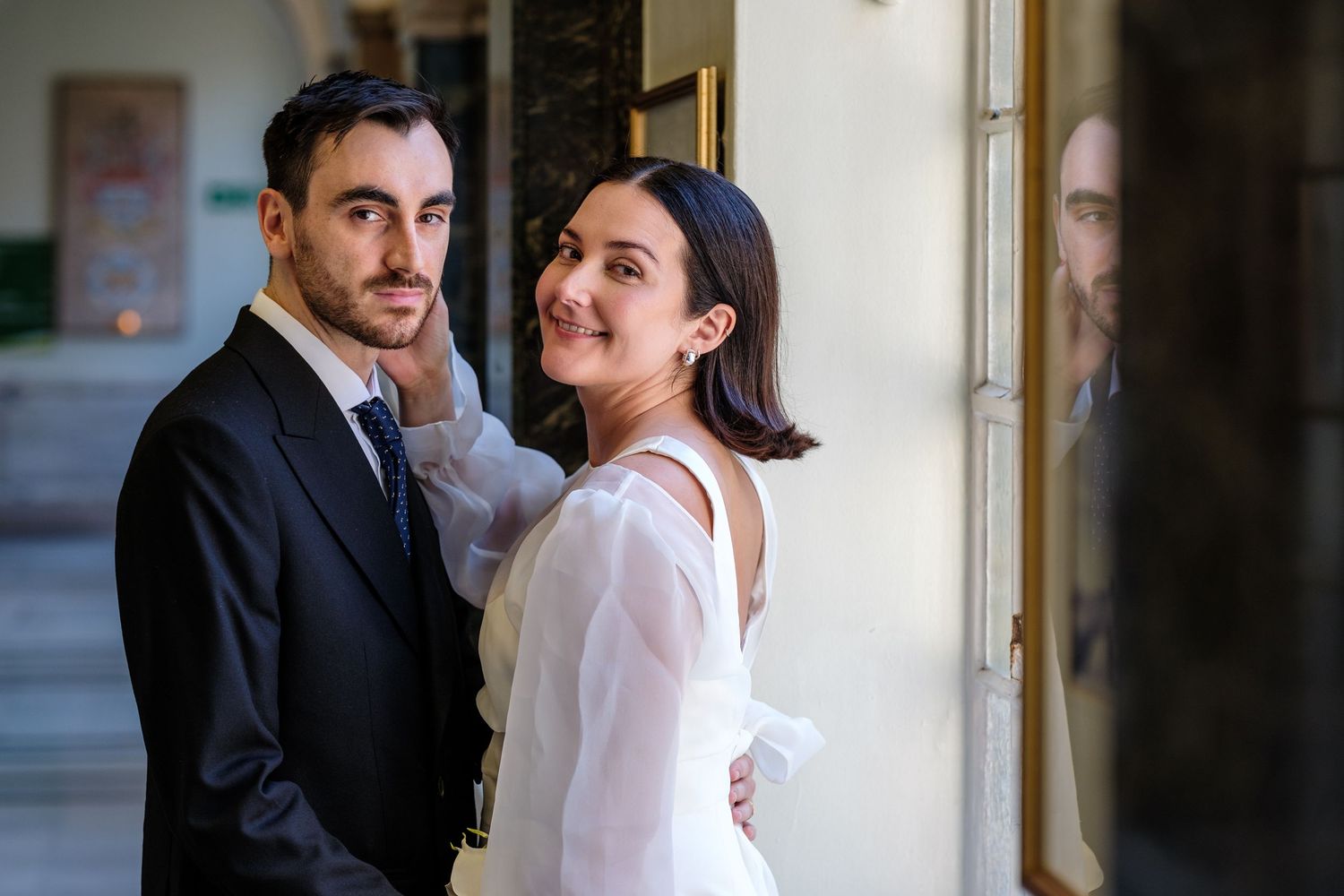 Elegant couple in wedding attire posing near ornate columns, bride in white dress adjusting grooms dark suit jacket.