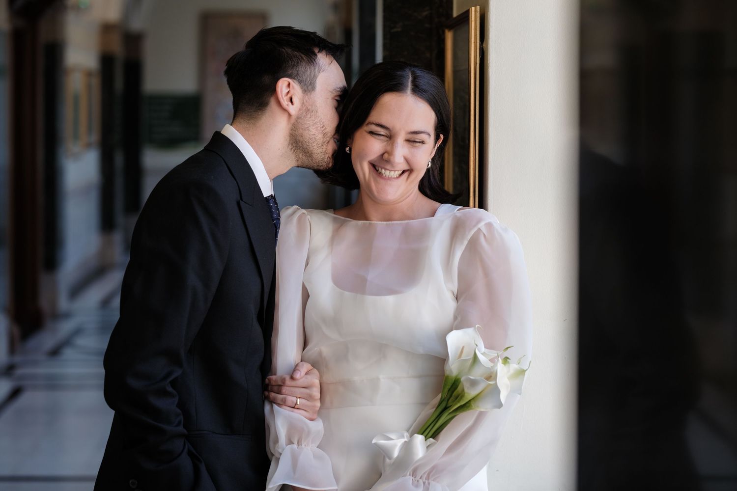 Bride in white gown holding calla lilies smiles as groom in black suit whispers in her ear during wedding day.