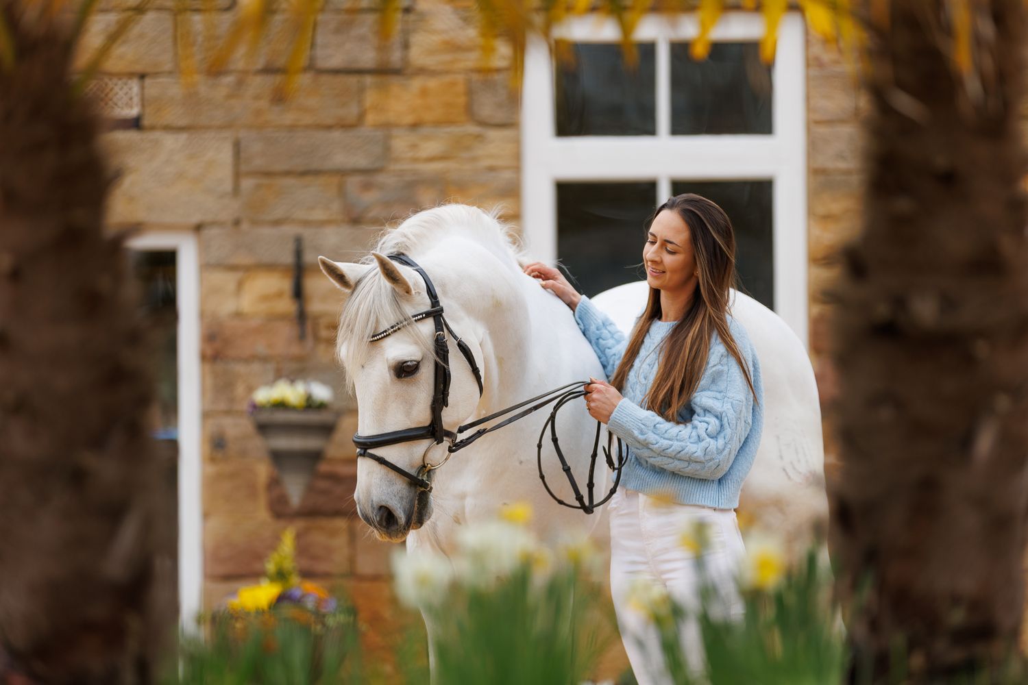 A girl stands between a white and brown horse at the entrance of a stone castle with a coat of arms.