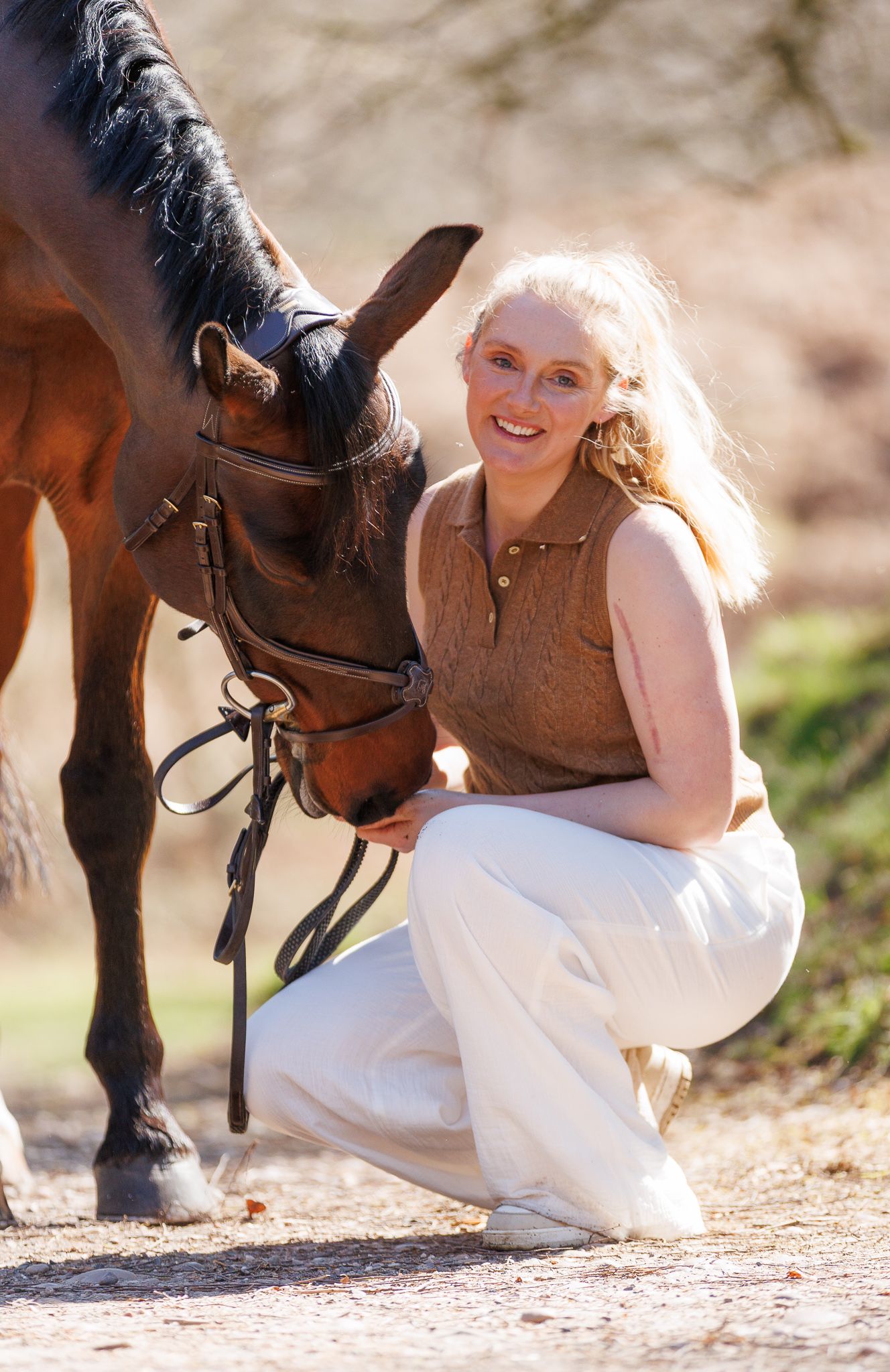 A brown horse and rider in a white sweater pose together against autumn foliage in soft natural lighting.
