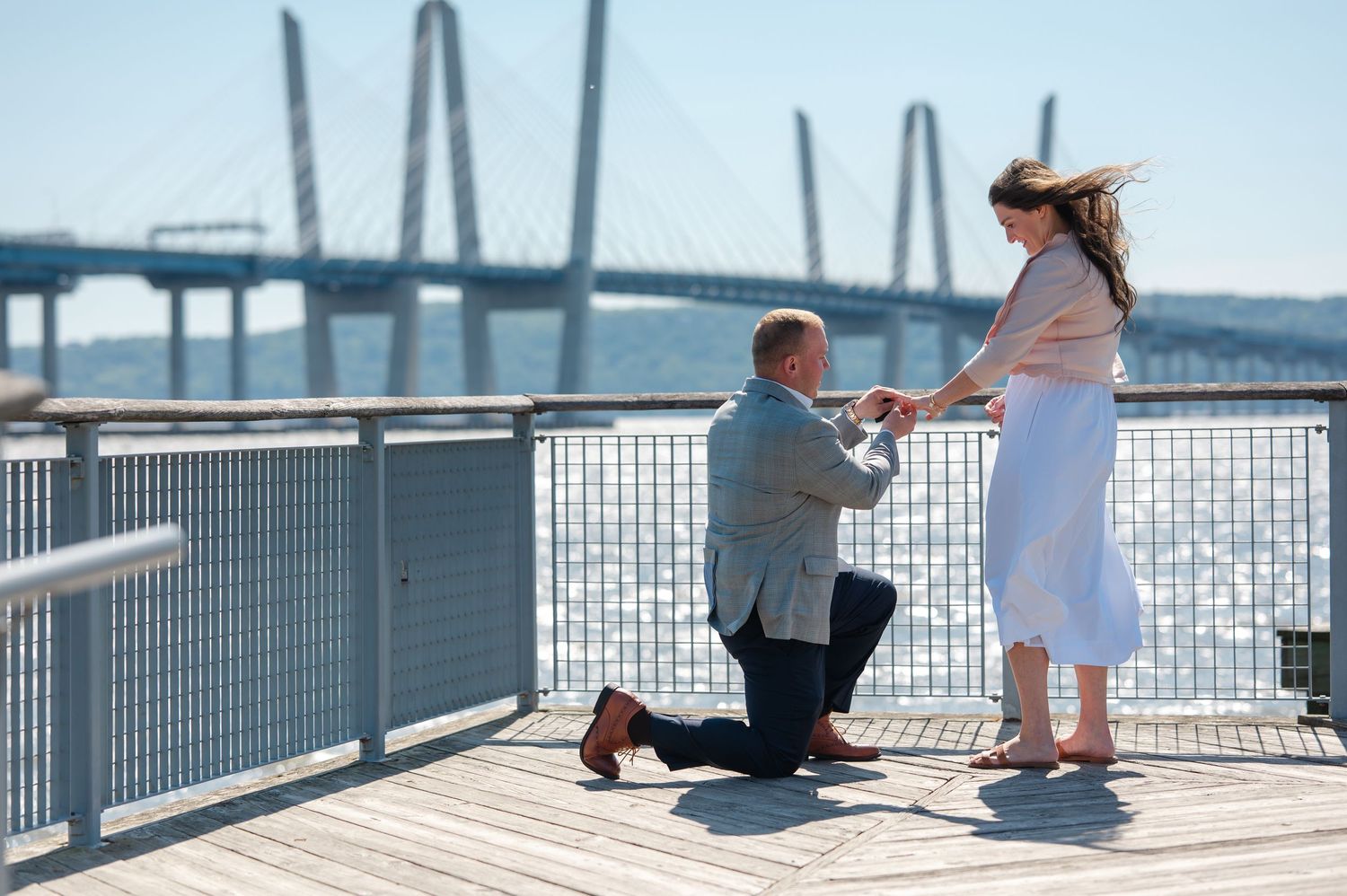 Man proposing on one knee to woman on waterfront deck with bridge in background on sunny day.