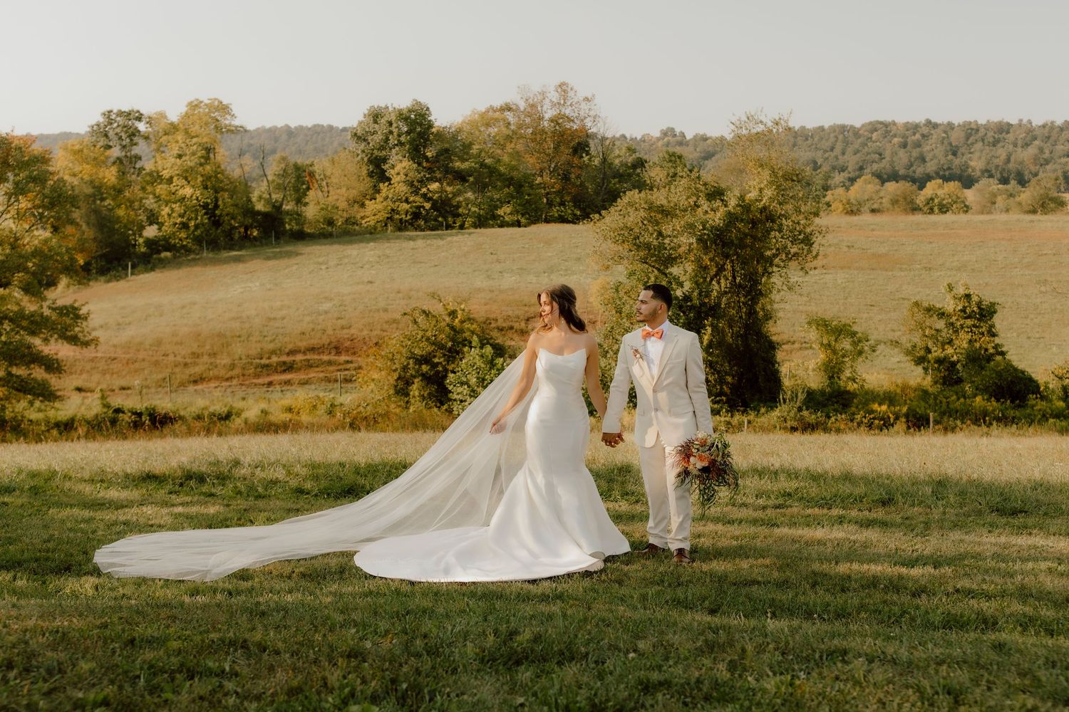 Bride and groom walk hand-in-hand across a sunlit green field with rolling hills and trees in the background.
