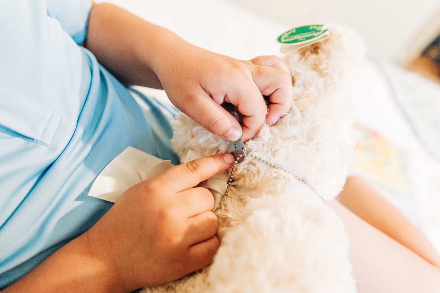 Child's hands pointing to a necklace on his teddy bear that says "DAD"