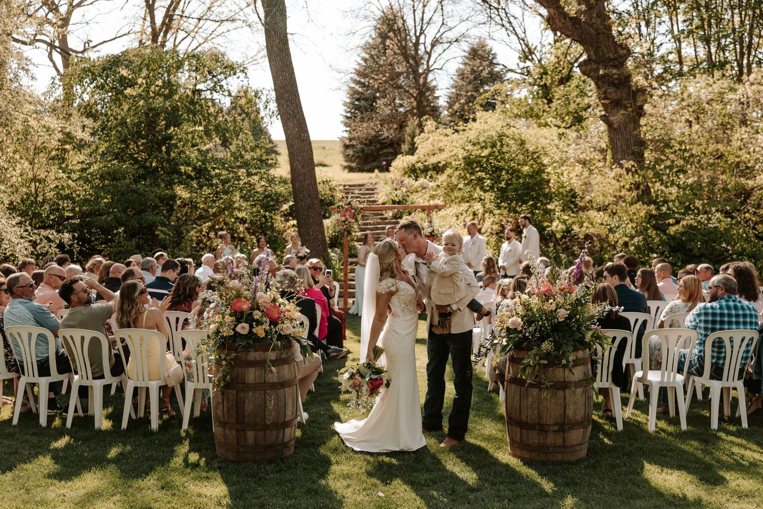 Smiling blonde bride in a flowing white boho wedding dress twirling outdoors in a lush green park setting.