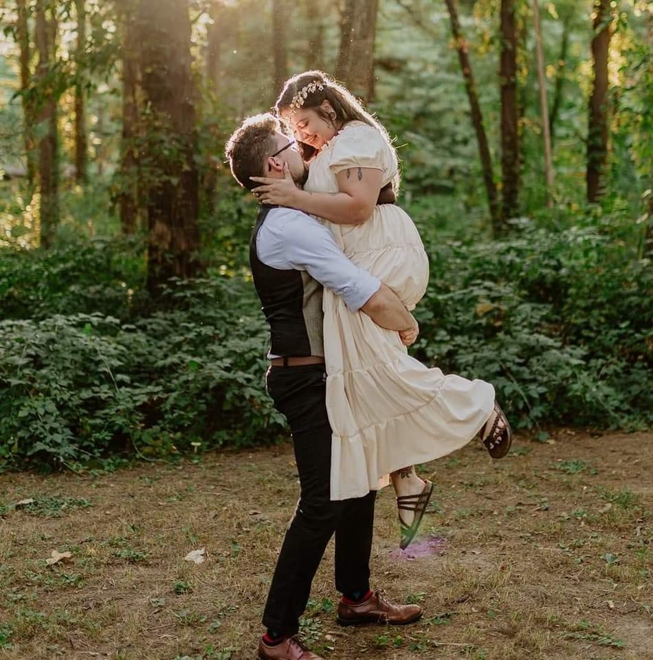A groom lifts his bride in a romantic kiss among lush green forest trees during an outdoor wedding.