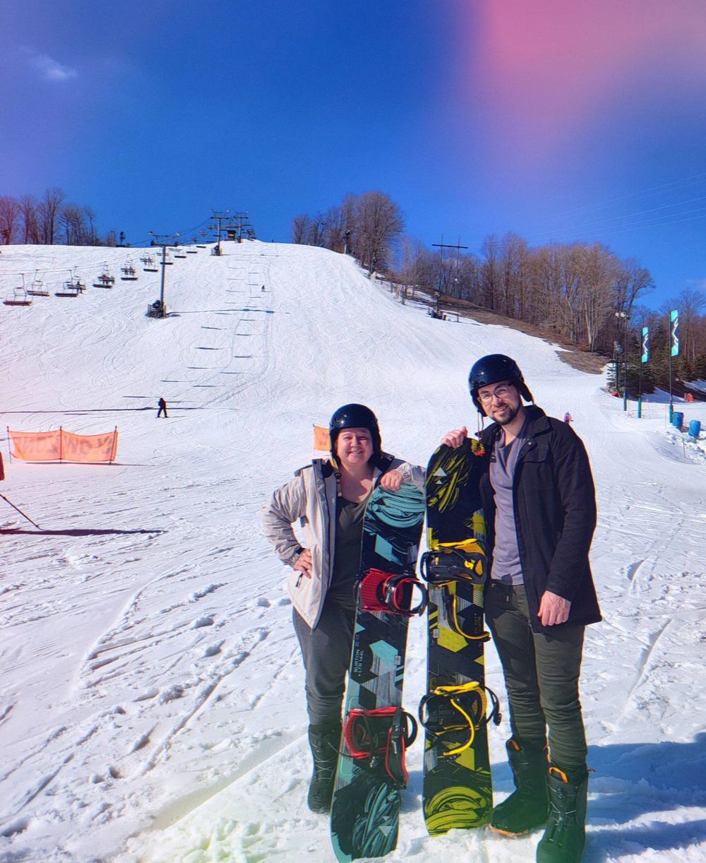 Two people posing with snowboards on a sunny ski slope with a chairlift and snowy hill in the background.