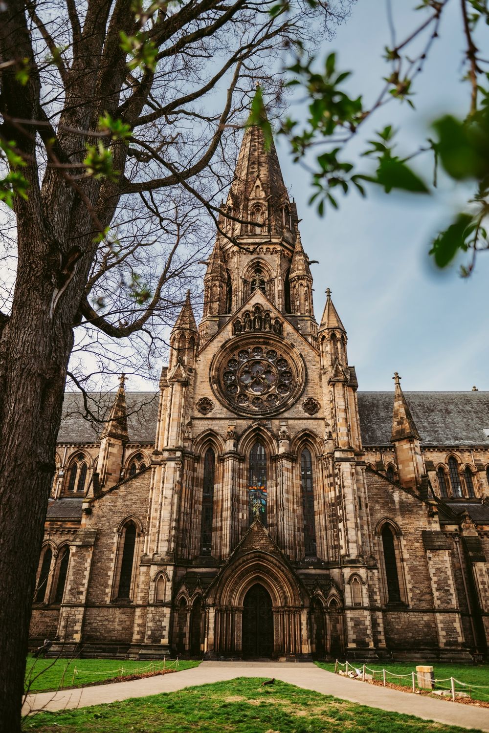 Gothic stone cathedral with tall spire, rose window, and arched entrance, framed by trees in a green courtyard.